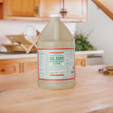A large bottle of Dr. Bronners Pure Castile Soap sits on a wooden kitchen counter. The background features wooden cabinets, a dish rack, and a plant, suggesting a cozy, eco friendly kitchen setting [1 Gallon]