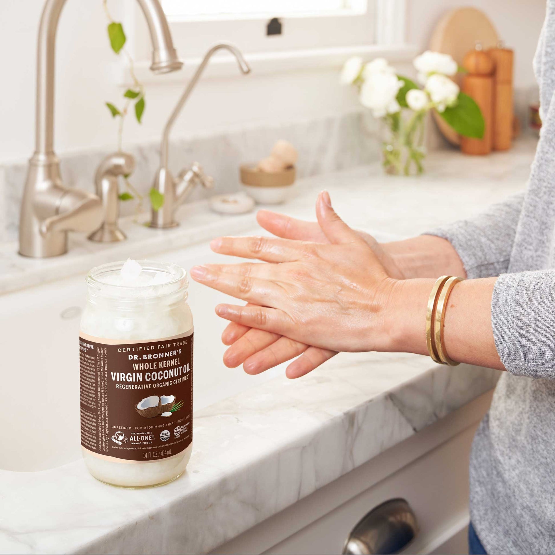 Hands apply Dr. Bronners Whole Kernel Virgin Coconut Oil in a modern kitchen. Labeled as certified fair trade and regenerative organic certified, the jar rests on a marble countertop near a stainless steel sink [14 oz]