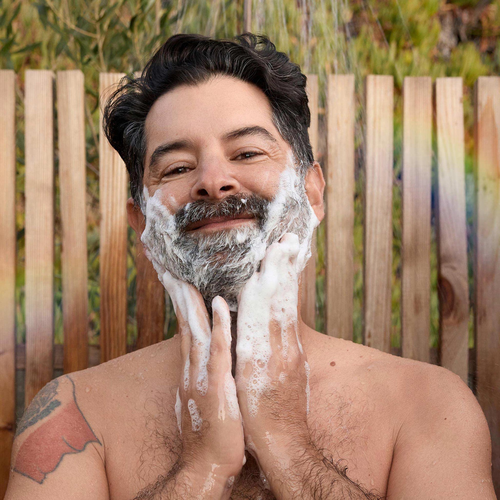 A man with a soapy beard and smile washes his face using dr bronners castile soap, standing outdoors against a wooden fence. Lush greenery surrounds the setting, creating a natural vibe [2 oz, 3.4 oz, 4 oz, 8 oz, 16 oz, 32 oz, 1/2 Gallon, 1 Gallon]